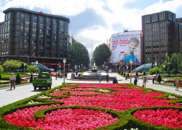 Люди идут мимо цветочных клумб, фонтана на площади Моюа